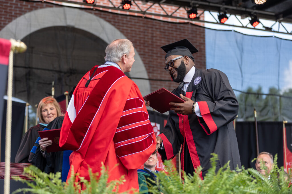 A graduate in a black gown and cap receives a diploma from a faculty member in red academic robes during an outdoor Professional Studies graduation ceremony. Other people are seated and smiling in the background.