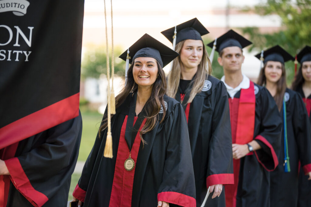 A group of graduates in black caps and gowns with red stoles stand in a line outdoors, smiling as they prepare to participate in a 00-Component graduation ceremony. A university banner is partially visible on the left.