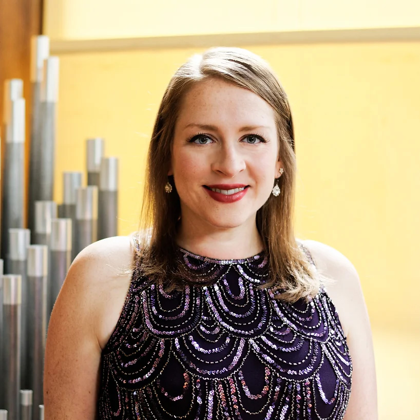 Amanda Mole, wearing a sleeveless, purple beaded dress and earrings, smiles at the camera against a backdrop of metallic tubes and a yellow wall—perhaps ready to perform at the McAfee Organ Concert.