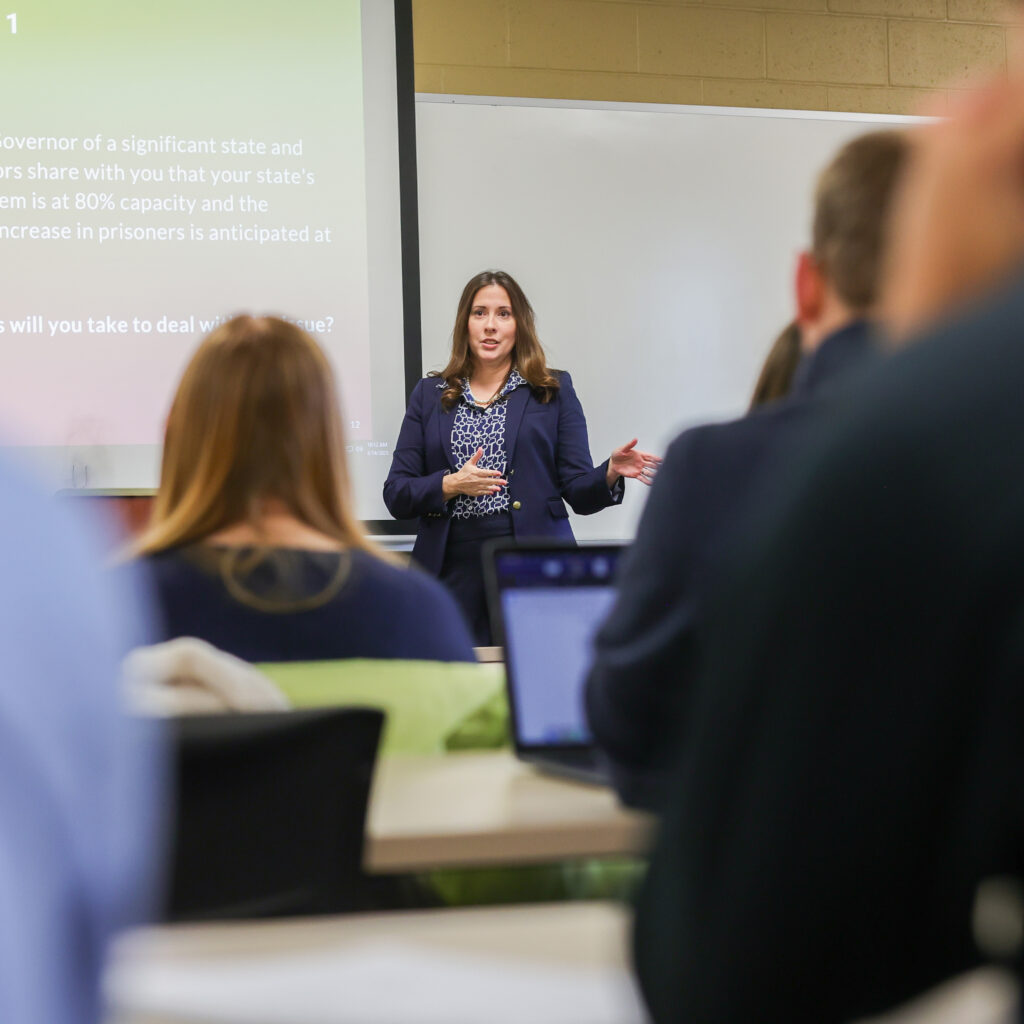 A woman in business attire stands at the front of a classroom giving a presentation to business students, while several seated people listen. There is a projection screen with text and a whiteboard behind her, reflecting the tri-school collaboration at McAfee School of Business.