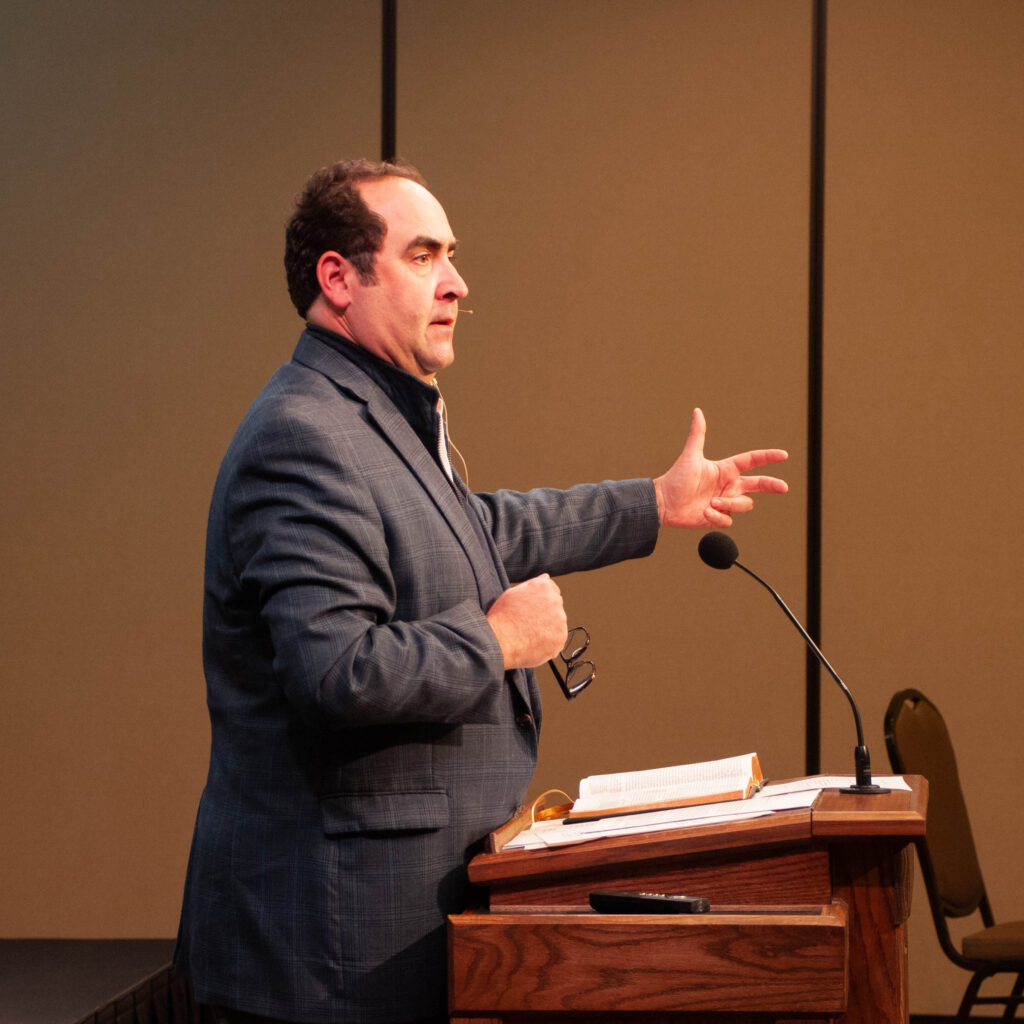 At the Ryan Center Bible Conference, Dr. Gignilliat, in a blue blazer, speaks at a podium with an open book—likely Ecclesiastes—gesturing with one hand and holding glasses in the other. A microphone, brown walls, and a chair are visible in the background.
