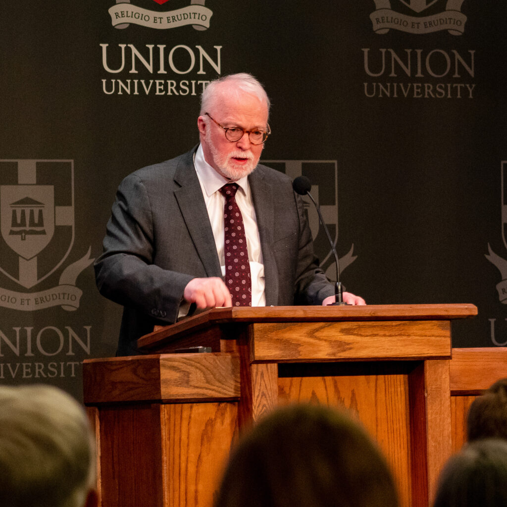 An older man with white hair and a beard speaks at a wooden podium with a microphone, in front of a backdrop that reads Union University with the school’s crest, during the David and Lanese Dockery Lectureship on Baptist Heritage. Audience members are visible in the foreground.