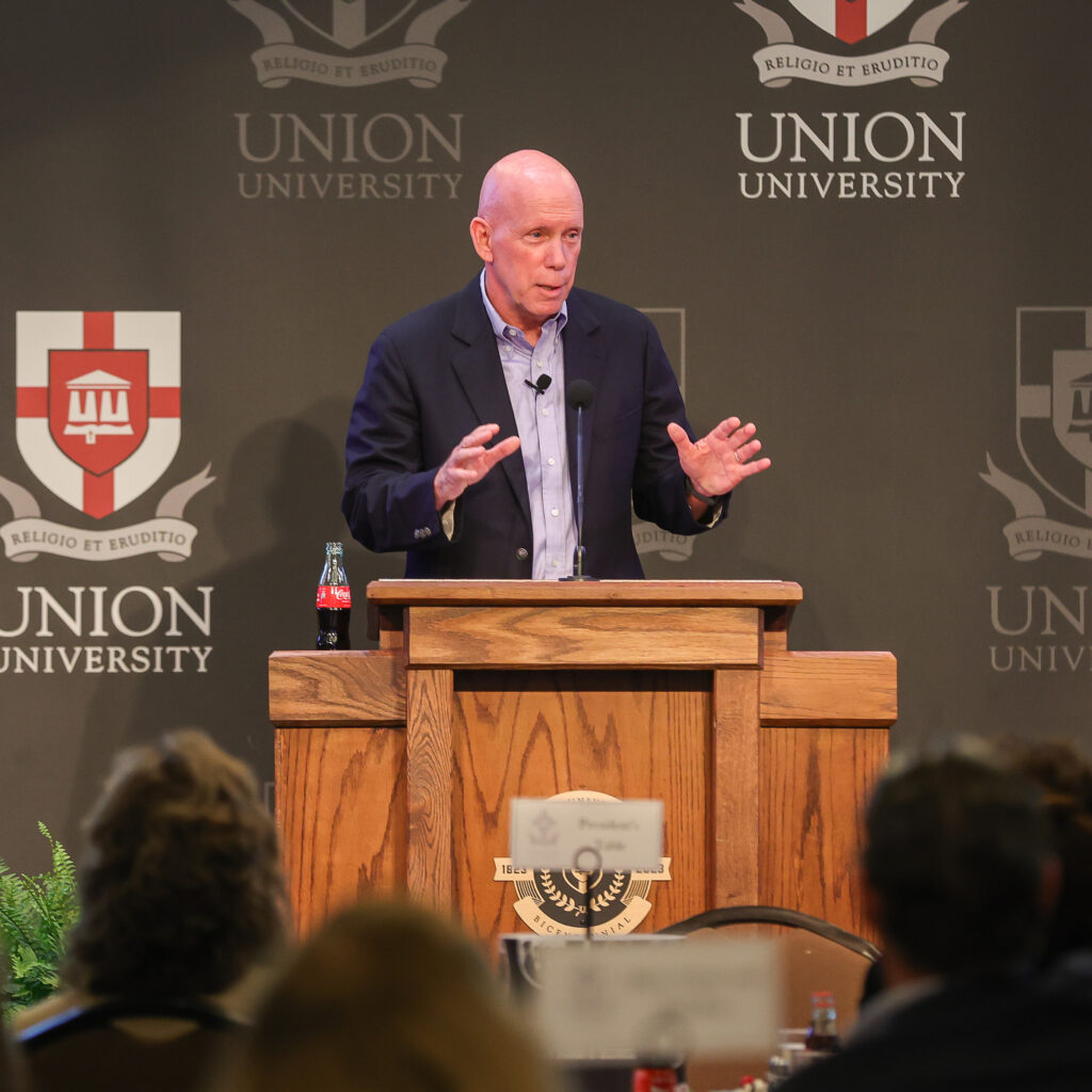 A bald man in a suit speaks at a wooden podium with a Coca-Cola bottle on it, in front of the Union University logo and motto, sharing insights about legacy and purpose. People are seated in the audience, listening intently.