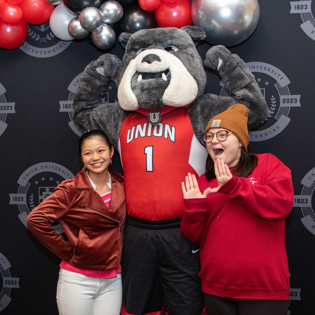 Two people pose with a gray bulldog mascot in a red Union jersey, standing before a Union University and Centennial 1823-2023 backdrop with balloons, celebrating the Union 200th anniversary.