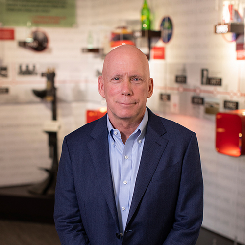 A bald man in a blue suit jacket and light blue shirt stands indoors at the Faith in the Marketplace luncheon, with display shelves and informational panels blurred behind him.