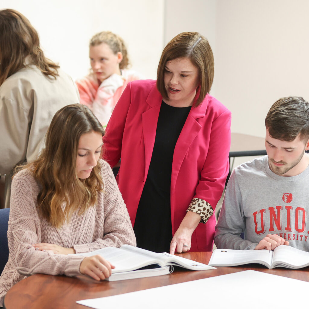 A woman in a bright pink blazer stands beside two students at a table, looking at open textbooks. In this School of Education classroom, others are visible in the background, reflecting the pursuit of a high score on the state report card.