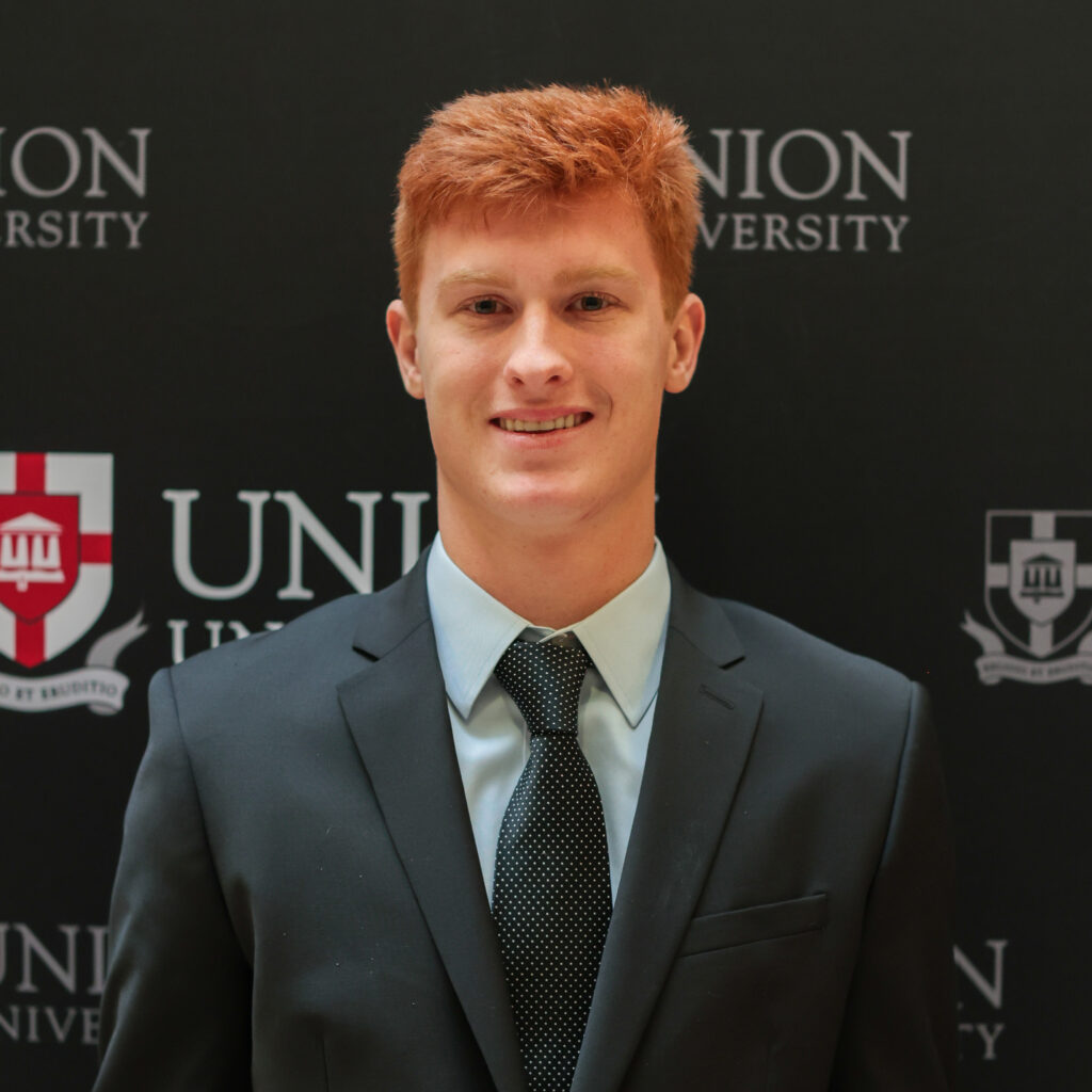 A young man with short red hair, Naranjo, wearing a dark suit, light blue shirt, and black tie stands in front of a Union University backdrop after receiving the Union top academic scholarship.