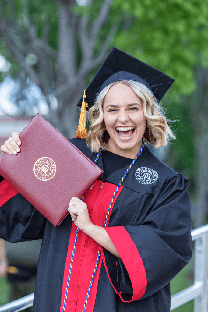 A person in a graduation cap and gown holding a diploma folder, smiling widely. The gown is black with red accents, and there is a green, leafy background outdoors.