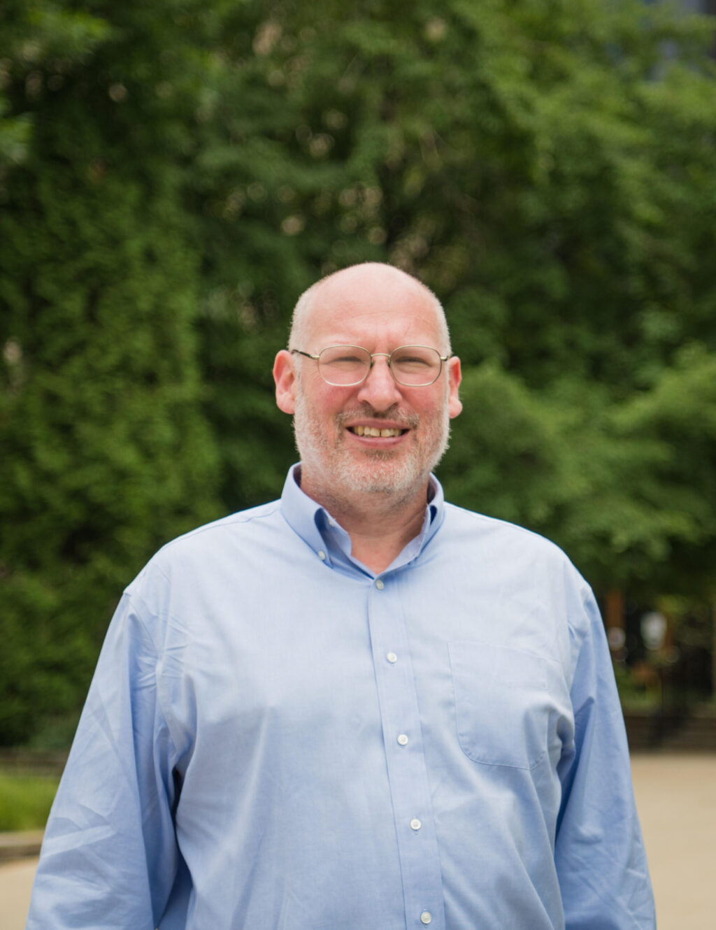 A middle-aged man with glasses and a bald head smiles while standing outdoors. He is wearing a light blue button-up shirt, and green trees are visible in the blurred background. This is Steven Pincus, who will be featured in the Carls-Schwerdfeger History Lectures in 2026.