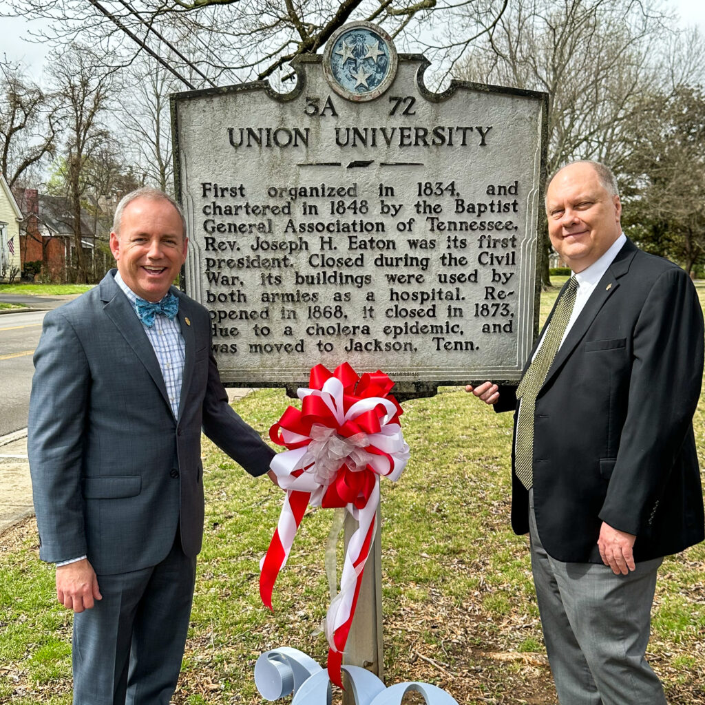 Two men in suits stand on either side of a historical marker for Union University in Murfreesboro. The marker, adorned with a red and white ribbon bow, stands near trees and a street visible in the background.