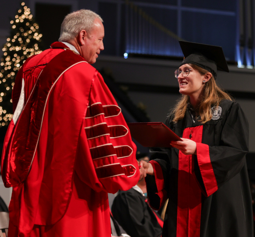During Union December commencement, a person in academic regalia hands a diploma to a graduate in cap and gown as they shake hands on stage, with a decorated tree visible in the background.