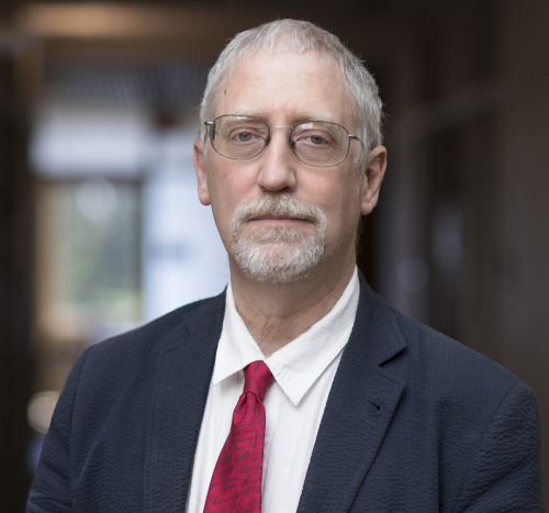 A middle-aged man with gray hair, a beard, and glasses wears a navy suit, white shirt, and red tie. The Carls-Schwerdfeger History lecturer for 2023 stands indoors with a blurred background and looks directly at the camera.