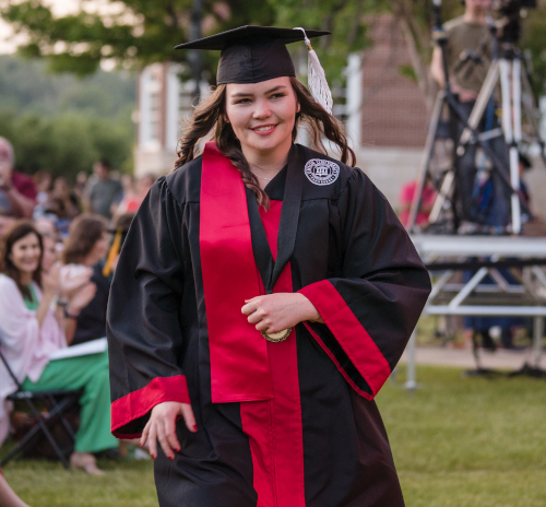 A graduate in a black cap and gown with a red stole walks outside during Union University's spring commencement ceremony. People are seated and clapping in the background, with green trees and a building visible.