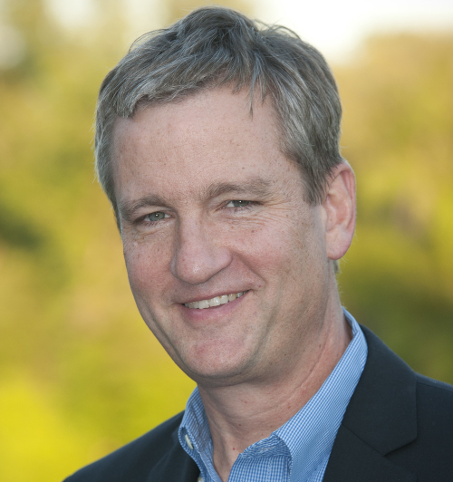 A man with short, light brown hair is wearing a blue collared shirt and dark blazer. He is smiling outdoors, with a blurred green and yellow background at the Faith in the Marketplace Union luncheon hosted by Barnhart.