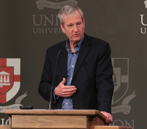 A man in a dark blazer and blue shirt stands behind a podium, speaking into a microphone. University crests and partial text—possibly mentioning Barnhart—are visible on the backdrop behind him.