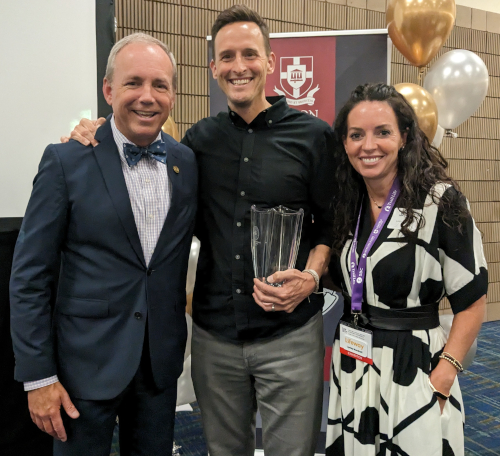 Three people stand indoors posing for a photo; the man in the center, Mandrell, holds a Dodd Award glass trophy. They are dressed in business or formal attire, with balloons and a Lifeway banner visible in the background.