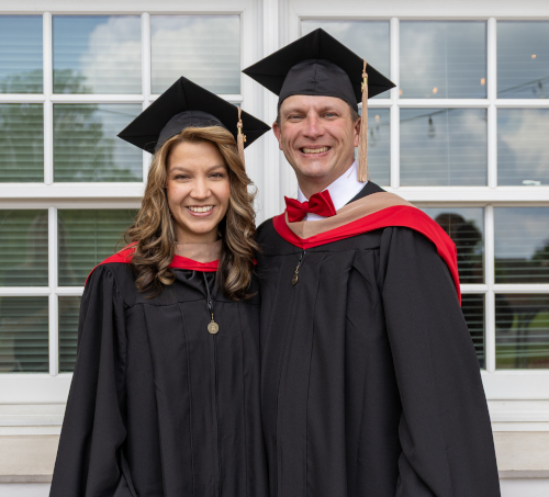 Two people wearing black graduation gowns and caps with red accents stand side by side, smiling, as they celebrate their father-daughter graduation in front of a window with white frames.