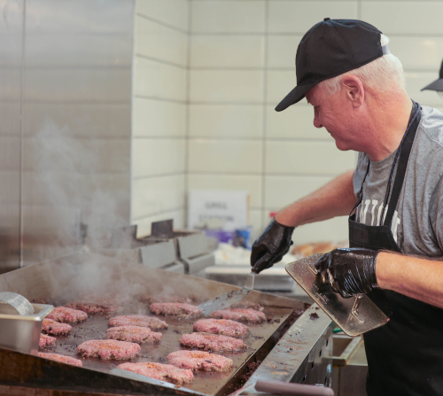 A person wearing a black hat, gloves, and apron cooks multiple beef patties on a large flat grill in The Lex’s commercial kitchen with tiled walls. Steam rises from the hot surface.