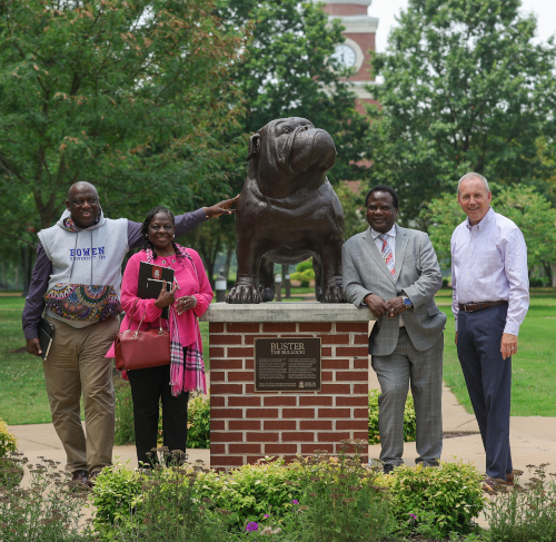Four adults stand outdoors next to a large bulldog statue on a brick pedestal with a plaque, smiling. Behind them are trees and Bowen University’s clock tower in Nigeria, symbolizing partnership and achievement.