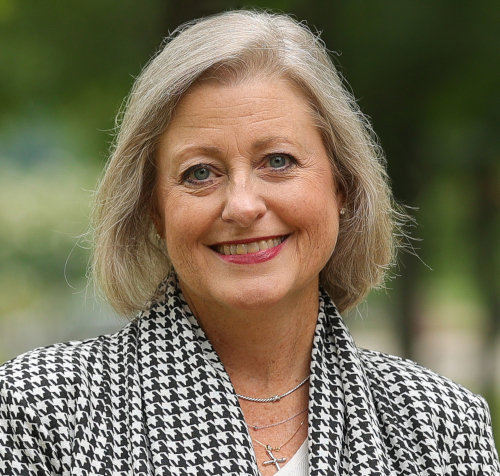 A woman with shoulder-length gray hair, believed to be Union SOAPS dean Emery, is smiling at the camera. She wears a black and white houndstooth jacket and a cross pendant necklace, standing outdoors surrounded by greenery.