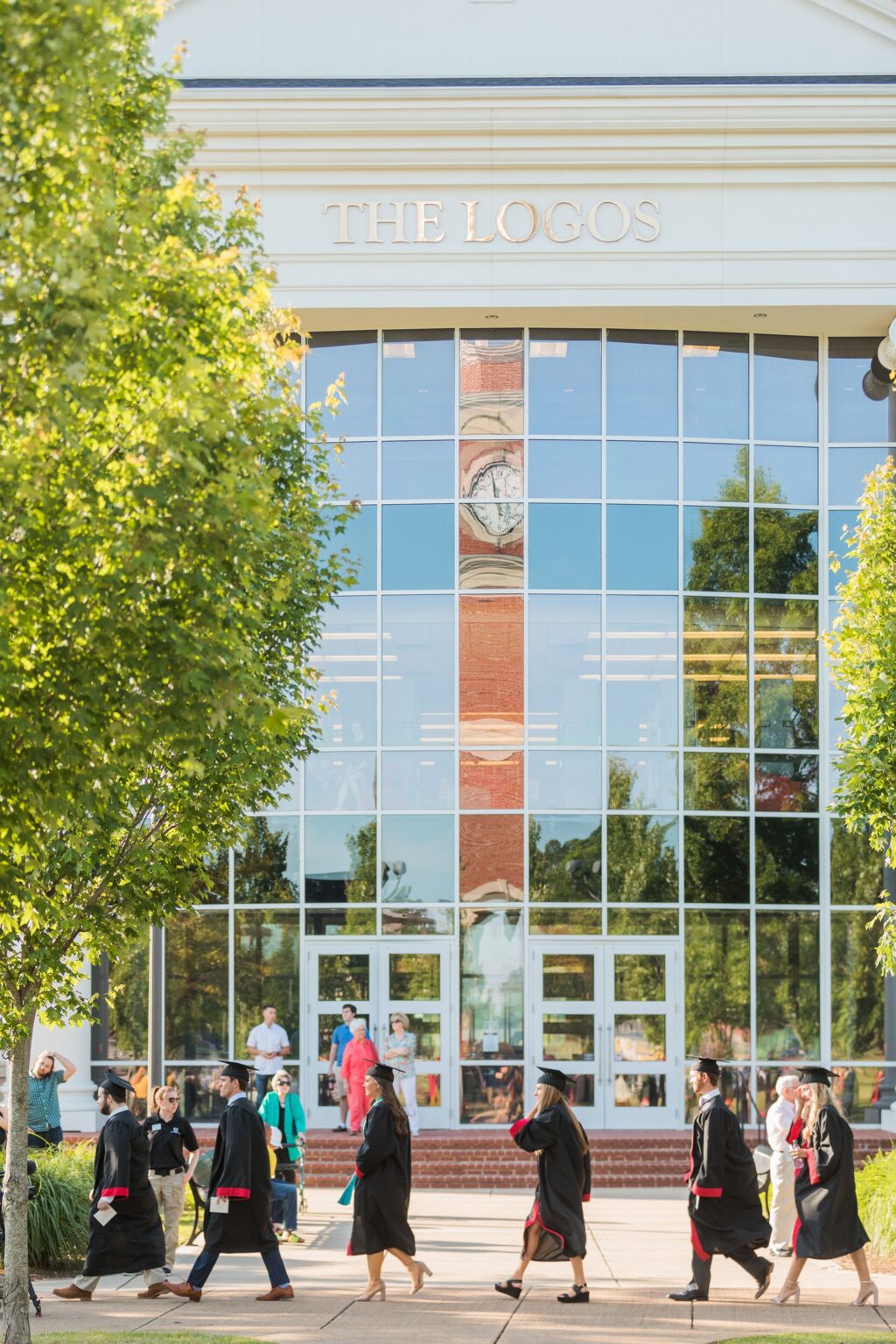 People in graduation gowns walk in front of The Logos, a building with large glass windows. Other Component members stand and talk near the entrance as green trees frame the scene.