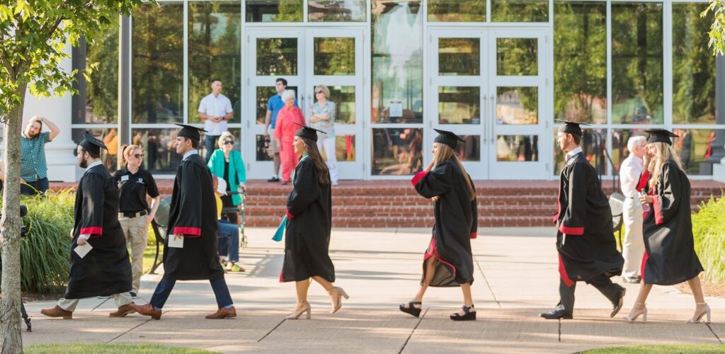 A group of graduates in caps and gowns walk in a line outside a building with glass doors, as an important component of their journey unfolds. Onlookers stand nearby, surrounded by trees and greenery along the walkway.