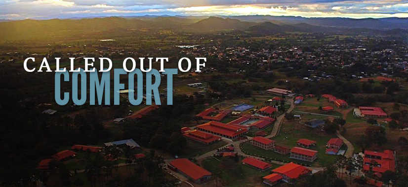 Aerial view of a campus with red-roofed buildings, surrounded by greenery and mountains under a cloudy sky. The phrase “Called out of Comfort” is boldly overlaid in large white and blue letters, echoing the inspiring theme.