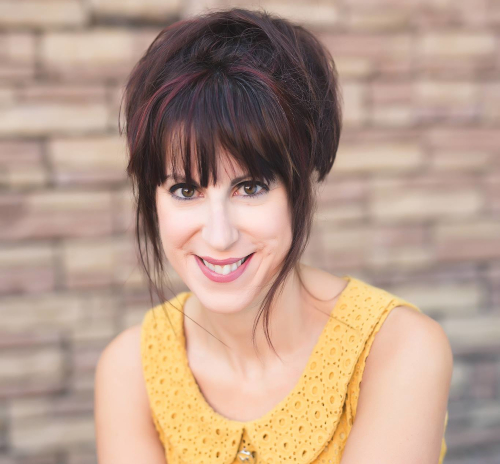 Bailey, a woman with shoulder-length dark hair and bangs, is smiling at the camera in a sleeveless yellow top. She stands before a blurred brick wall background at the Feb. 29 Creative Writing Workshop.
