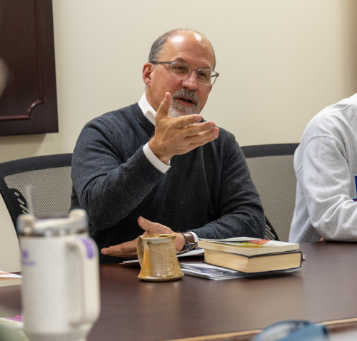 A man with glasses and a gray sweater, possibly a Classics major at Union University, gestures while speaking at a table. In front of him are a book, a mug, papers, and a water bottle. Two others sit partially visible beside him.