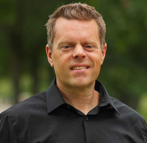 A man with short light brown hair, wearing a black collared shirt, stands outdoors in front of a blurred green background. He is looking at the camera with a neutral expression, representing the McAfee School of Business at Union.