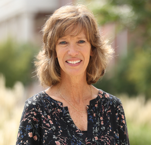 A woman with shoulder-length light brown hair smiles outdoors. She is wearing a black floral blouse and a thin gold necklace, representing business leadership at Union's McAfee School of Business, with greenery and a blurred building in the background.