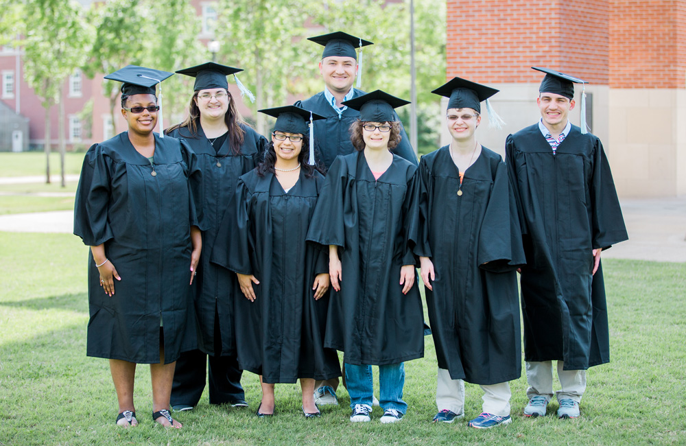 A group of seven students wearing graduation caps and gowns stand together on a lawn, smiling at the camera. Celebrating this milestone in student life, they pose with trees and brick buildings visible in the background.
