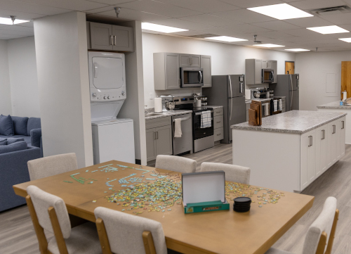 A modern communal kitchen at the EDGE facility features gray cabinets, stainless steel appliances, a stacked washer and dryer, and a wooden table with an unfinished puzzle. A blue sofa is visible in the background—perfect for Union gatherings on Jan. 30.