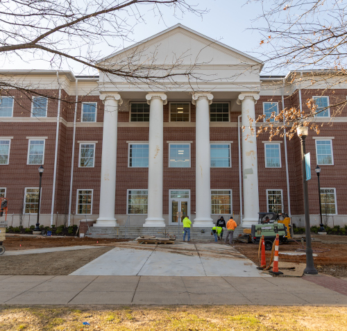 A three-story brick building with four large white columns at the entrance. Several construction workers and equipment are visible in front of The Ethos Forum, and the landscaping appears unfinished. Leafless tree branches frame the scene.