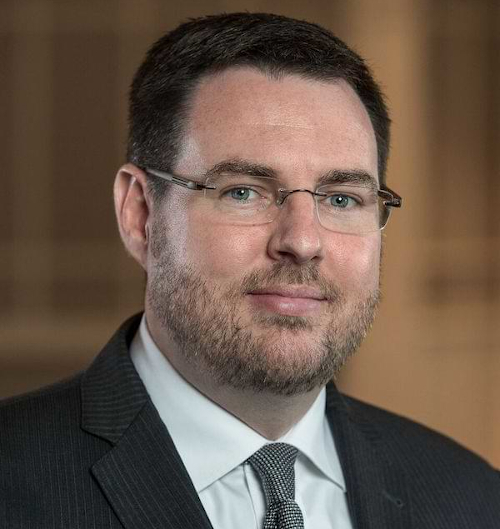 A man with short dark hair, glasses, and a trimmed beard is wearing a pinstripe suit, white shirt, and patterned tie, looking at the camera with a neutral expression. The blurred beige and brown background hints at BWA’s Brown setting.