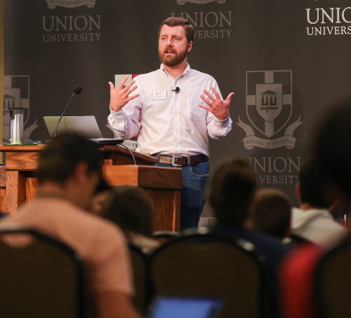 A man stands and gestures with both hands while speaking at a podium in front of an audience at the youth apologetics conference. The background displays the Union University logo and name on banners.