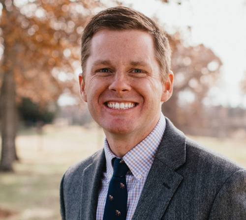 A man with short light brown hair, wearing a gray suit jacket, checkered shirt, and navy tie, is smiling outdoors with trees and blurred foliage in the background at the Dockery Lectures on March 20.