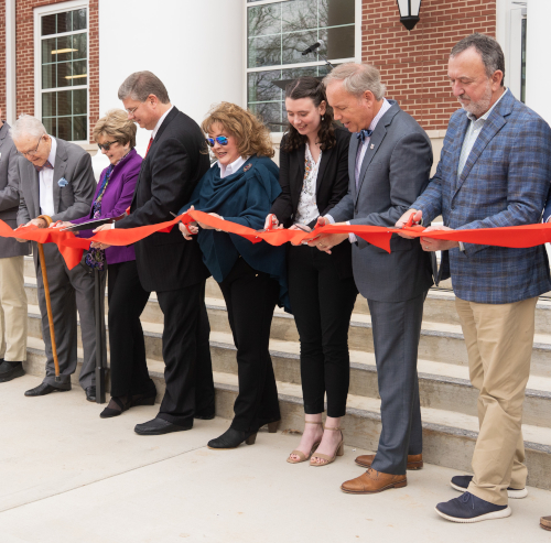 Seven adults stand in a row cutting a red ribbon with large scissors during The Ethos Forum’s outdoor ribbon-cutting ceremony, celebrating the Union building’s opening with brick walls and white columns as the backdrop.