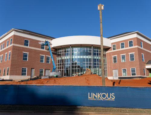 A modern brick building under construction with large glass windows, a curved entrance, and a blue safety fence labeled LINKOUS in front. A lift is parked nearby as The Ethos Forum academic hub takes shape, with dirt piled in front.