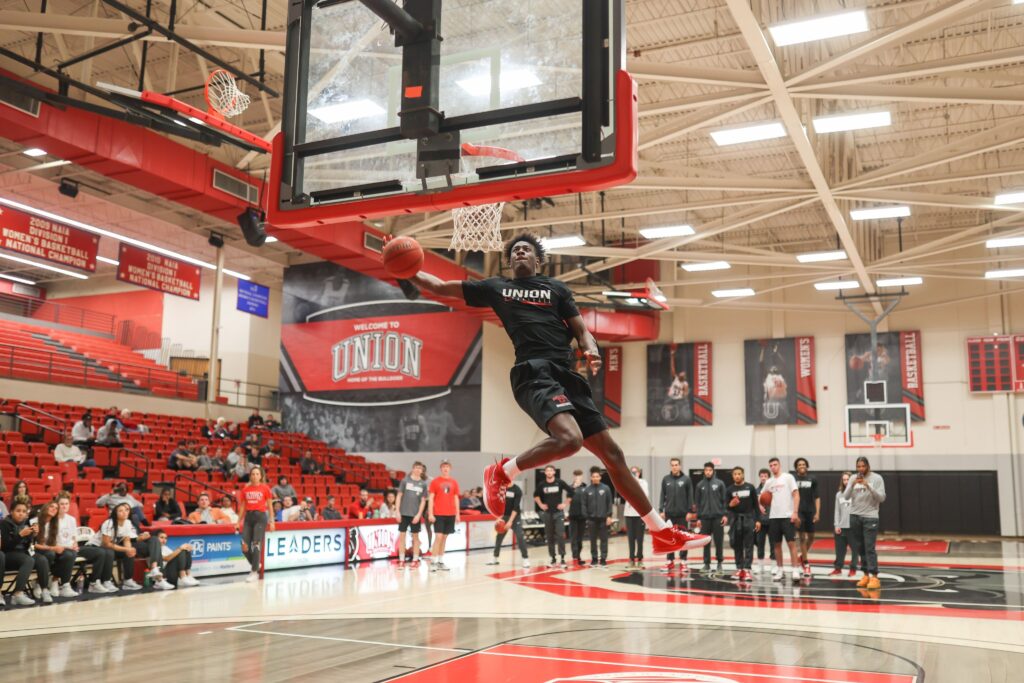 A basketball player in a black Union shirt jumps toward the hoop for a dunk in a gymnasium, with spectators and other players watching from the sidelines. The gym has red seats and banners on the walls.