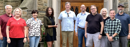 Ten adults stand in a row outside a stone building with a wooden door, posing for a group photo at a faculty mentoring program retreat. Most are casually dressed and smiling, with greenery visible next to the doorway.