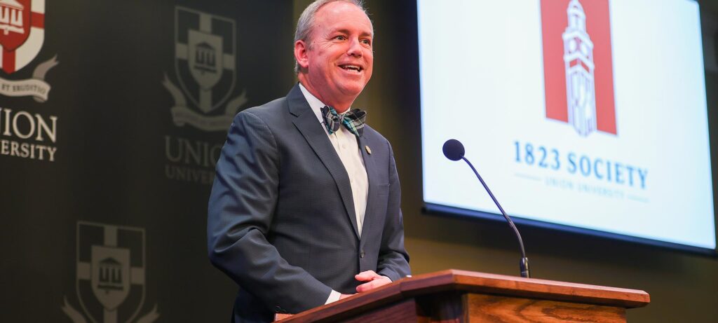 A man in a suit and bow tie stands at a wooden podium, inducts new members into the 1823 Society. Behind him are Union University banners and a screen displaying the Union University logo and 1823 Society.