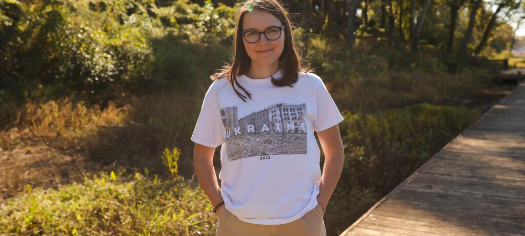 Anna Slabetska, wearing glasses and a white Ukraine 2022 t-shirt, stands alive and vibrant outdoors on a wooden path, surrounded by greenery and sunlight.