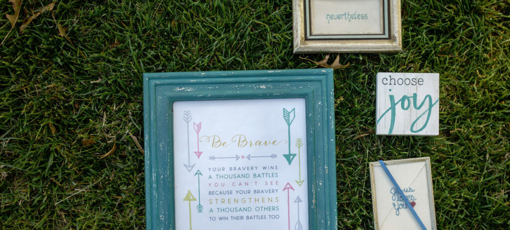 Four framed and canvas signs are arranged on grass. The largest sign, inspired by Beth Madison, says, “Be Brave. Your bravery wins a thousand battles you can’t see—strengthening a thousand others in their own struggles with chronic illness.”.