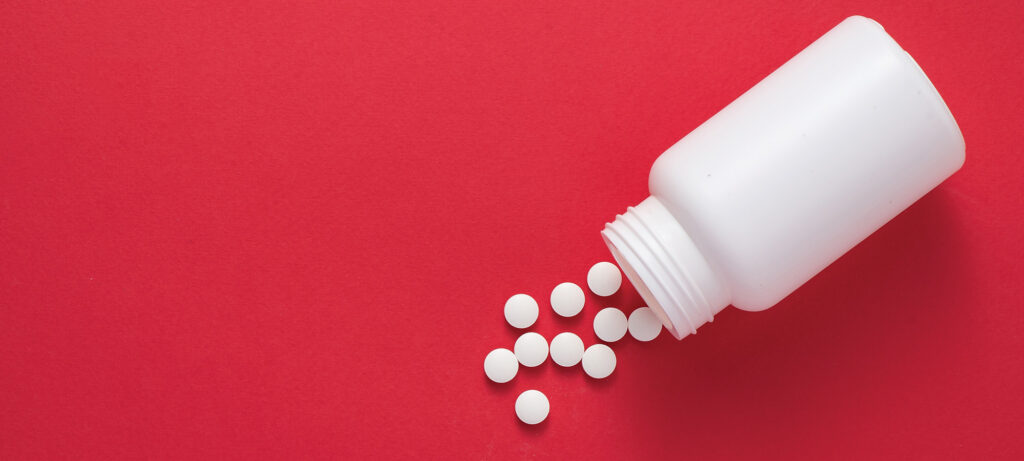 A white plastic pill bottle tipped over on its side with several round white tablets spilled out, all on a solid red background—evoking the urgent realities of the opioid crisis.