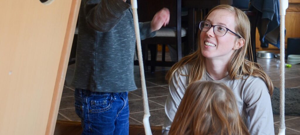 A stay-at-home mom with glasses, Chelsey Kurzhal, sits on a tiled floor, smiling and looking up at two children who are standing nearby. The children are partially out of frame, surrounded by household objects in the background.