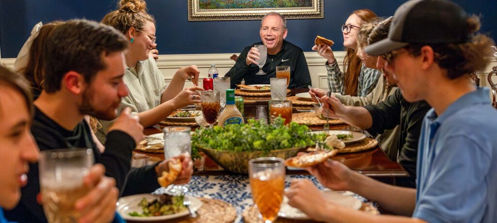 A group of people sit around a dining table set with plates, glasses, salad, and drinks. They are eating, drinking, and talking as Dub Oliver, at the head of the table, holds up a glass and smiles—celebrating together after running the race.