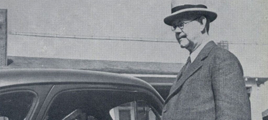 A man in a suit, glasses, and a hat stands next to a car, echoing a Moses figure amid the Union's wilderness wandering. The background reveals part of a building and a power line in this black-and-white image.