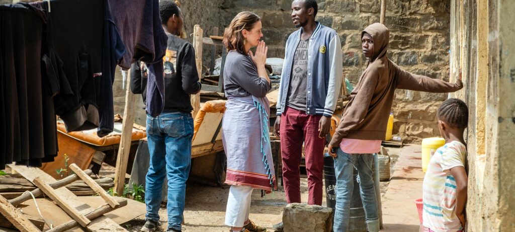 A group of five people, including Kristen Lowry and four men, stand and talk in an outdoor area with scattered furniture and laundry hanging nearby, next to a stone wall, lending their voice for the vulnerable in the community.