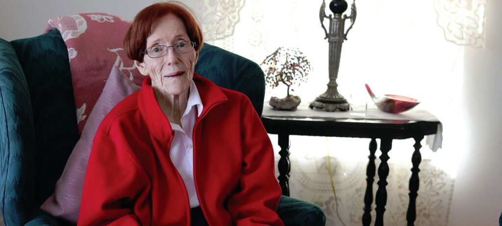 An elderly woman, reminiscent of a Female Senior Administrator, with short red hair and glasses sits on a green armchair in a cozy room. A table behind her holds a lamp, small plant, and decorative bowl, framed by lace curtains in the background.